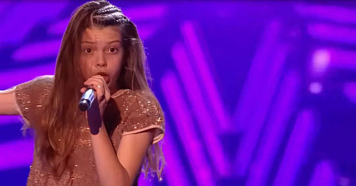 A person with long hair, wearing a sparkly top, sings into a microphone on a stage with purple lighting.