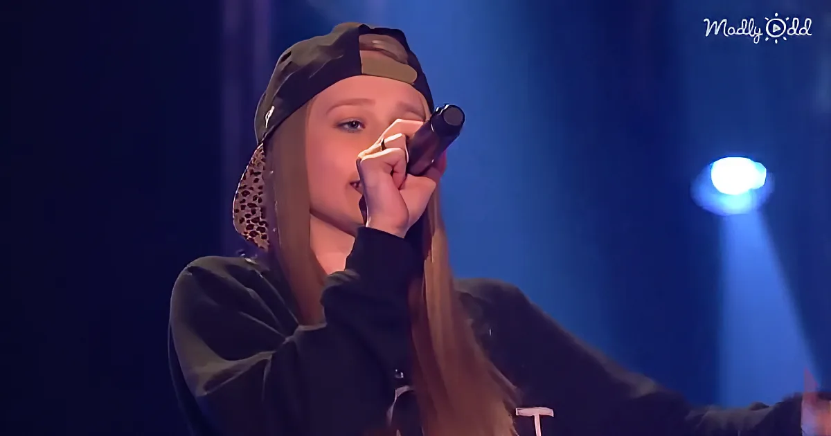A person wearing a cap and hoodie holds a microphone, performing on stage with blue lighting.