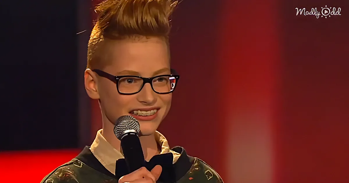 A person with a spiky hairstyle and glasses holds a microphone, smiling, against a red background.