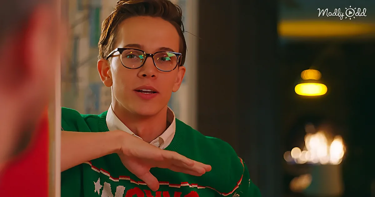 A person in a green sweater with a white shirt underneath, making a hand gesture, wearing glasses, and standing in a...