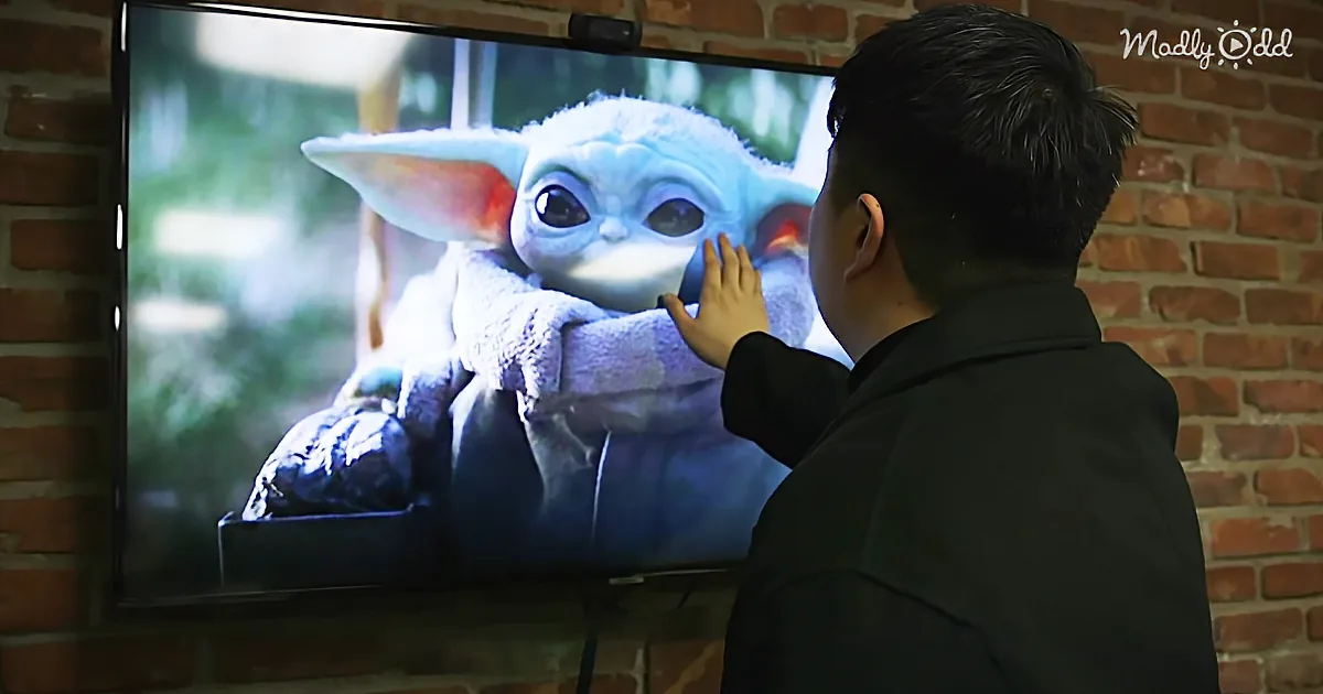 A boy reaches out to touch Baby Yoda on a TV screen in a brick-walled room.