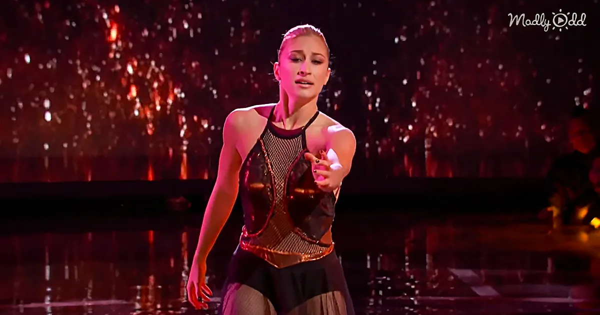 A figure skater in a black and gold outfit performs on a dark stage with red lighting and sparkling effects.