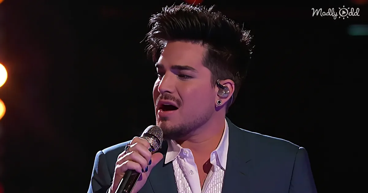A man in a suit sings into a microphone, wearing earpieces and a ring, with a dark background.