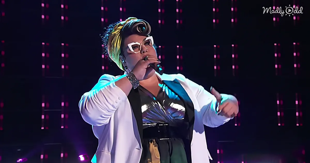 A performer in a white blazer and black outfit sings into a microphone on stage with a glowing background.