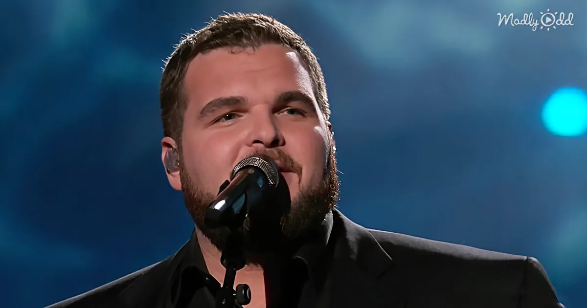 A man with a beard and short hair sings into a microphone, wearing a black suit, against a blue background.