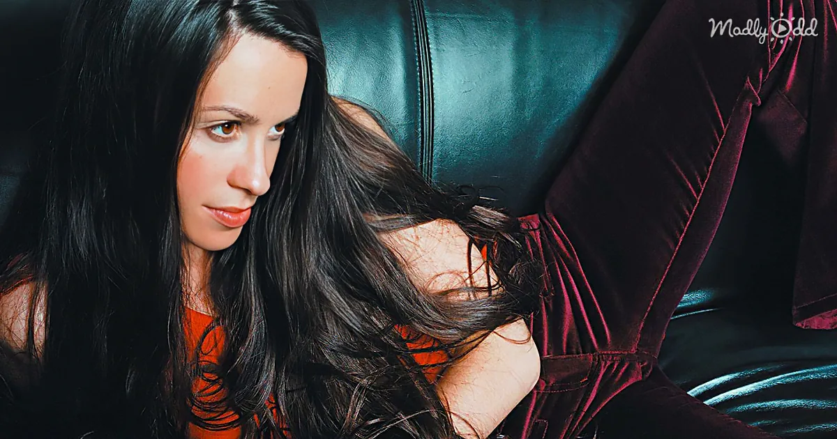 A woman with long dark hair in an orange dress leans on a black leather couch.