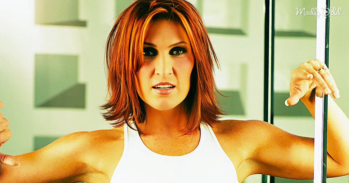 A woman with red hair in a white tank top flexes her muscles, holding a pole.