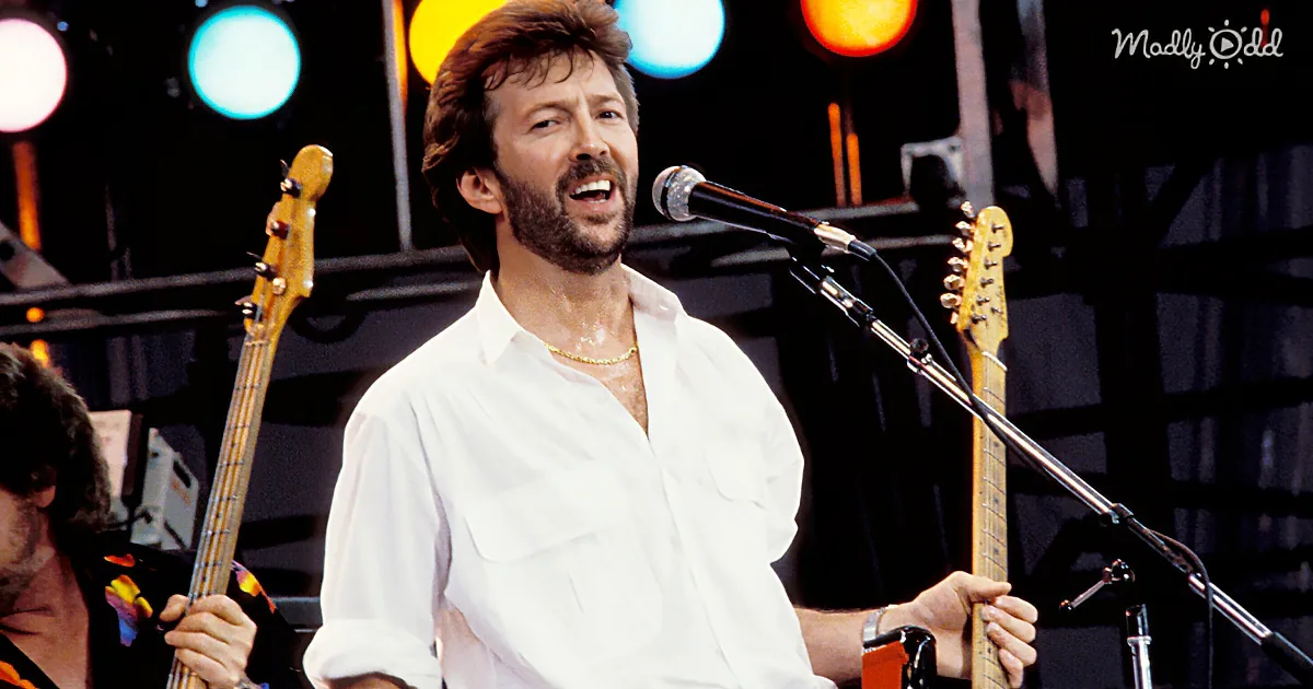 A man in a white shirt sings into a microphone, holding a guitar, with a band and stage lights in the background.