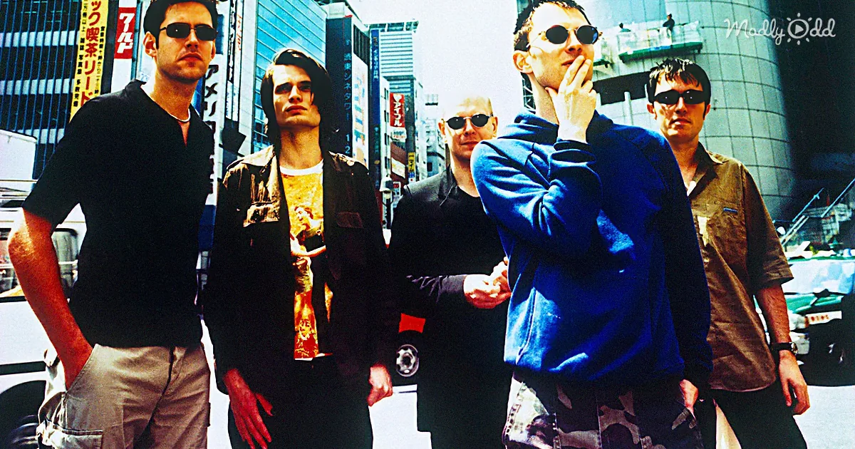 A group of five men stands on a city street, dressed casually, with one smoking and wearing sunglasses.