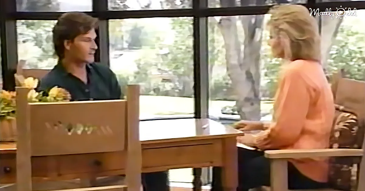 A man and woman are seated at a table, engaged in conversation in a room with large windows.