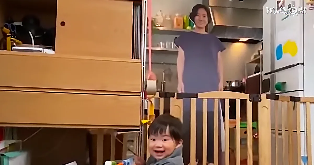 A child is smiling in a crib, with a woman standing behind a railing in a kitchen.