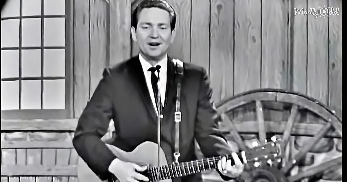 A man in a suit plays guitar and sings in front of a wooden backdrop.