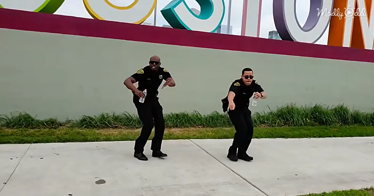 Two police officers in uniform dance on a sidewalk in front of a large colorful sign.
