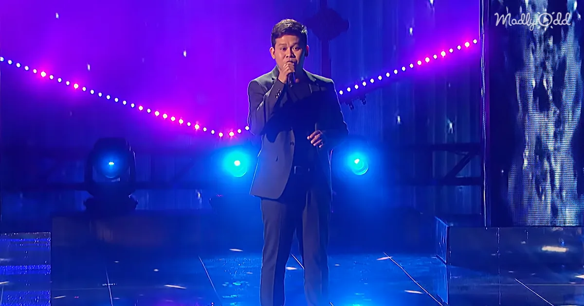 A man in a suit sings into a microphone on a stage with purple and blue lights.