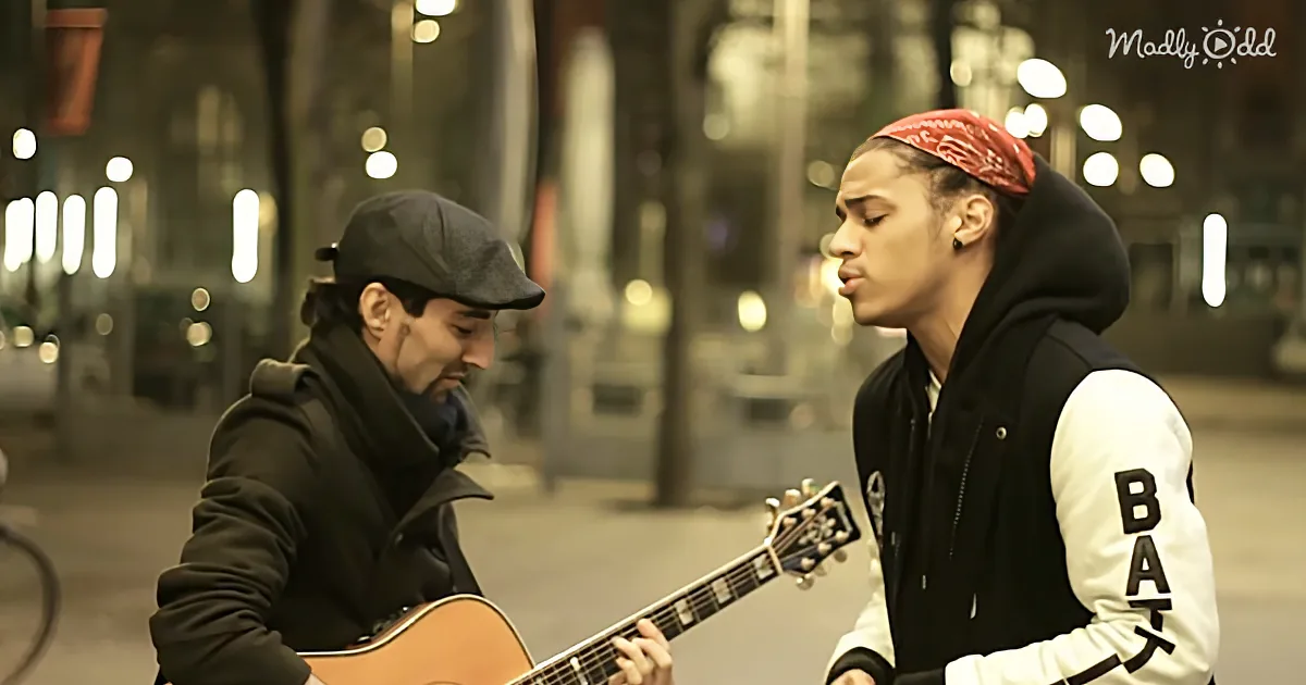 A man in a black jacket plays guitar while another in a red bandana listens intently on a city street at night.