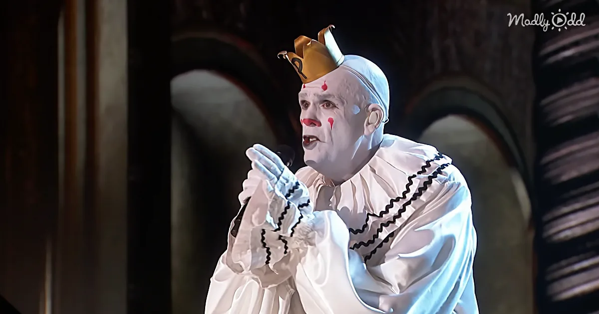 A person in a white clown costume with a crown and red nose is holding a microphone.
