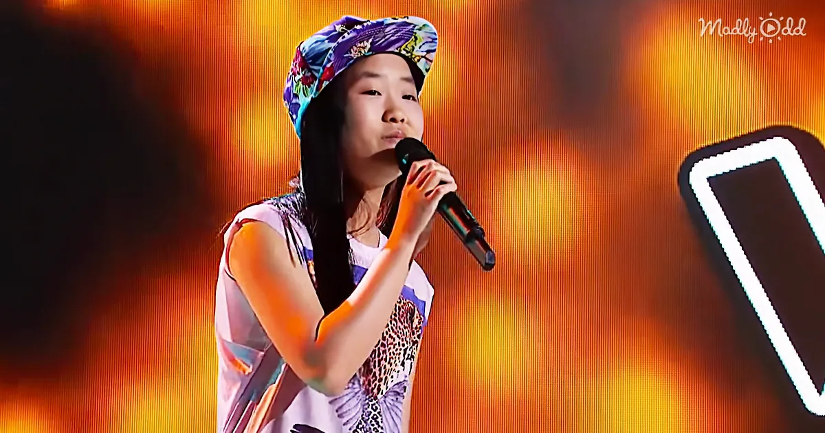 A young person wearing a colorful hat and patterned top sings into a microphone on stage with an orange backdrop.