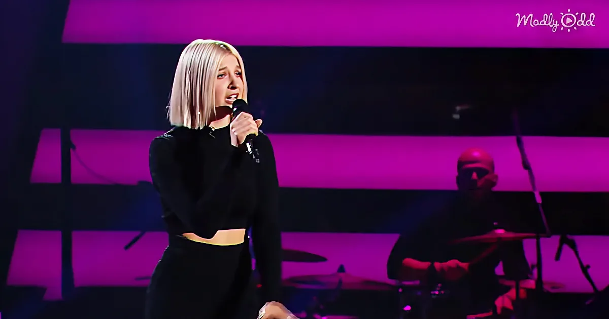 A woman with short blonde hair sings into a microphone on stage, with a drummer in the background and purple lighting.