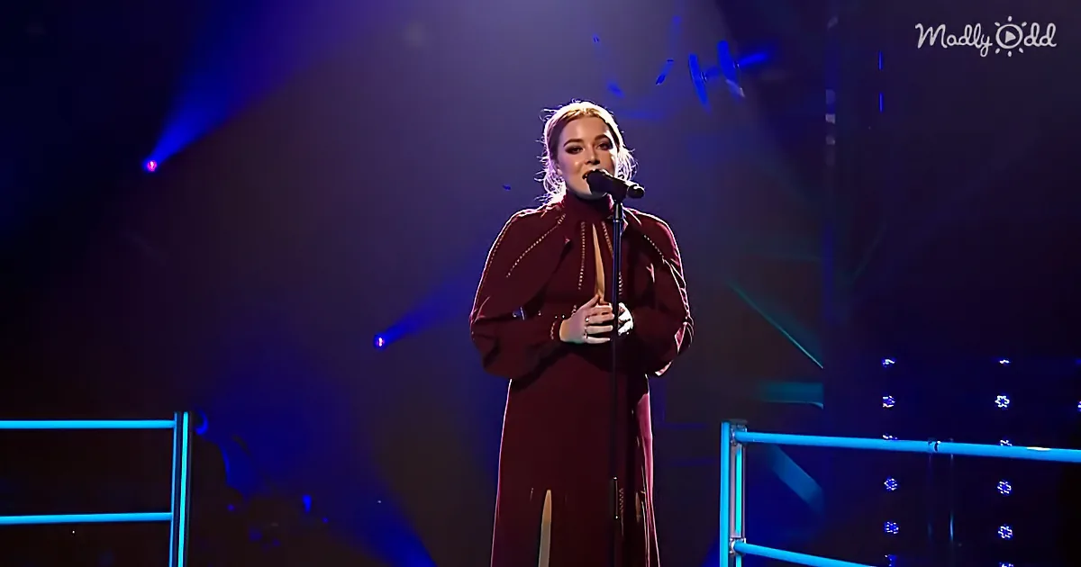 A woman in a maroon outfit sings into a microphone on a stage with blue lighting.