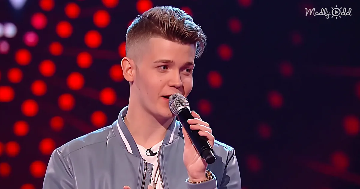 A young man in a light jacket holds a microphone, singing on stage with red lights in the background.