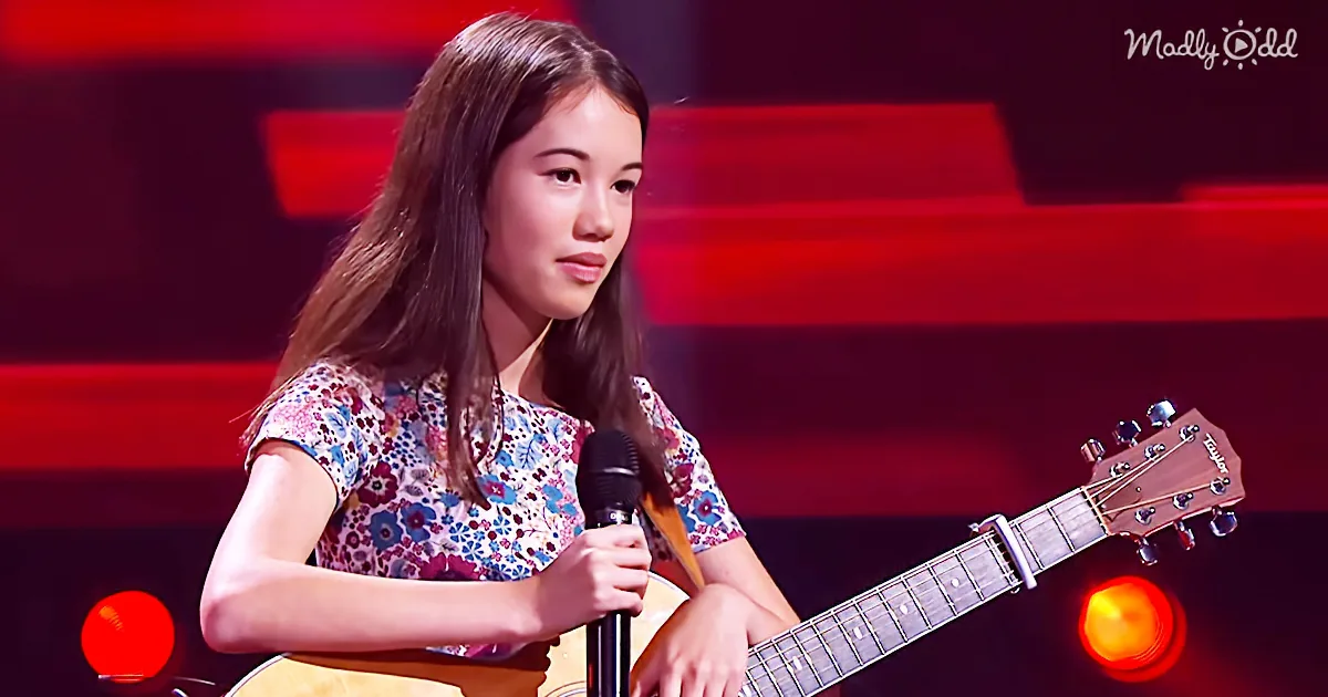 A young woman with long hair plays a guitar and sings into a microphone on stage.