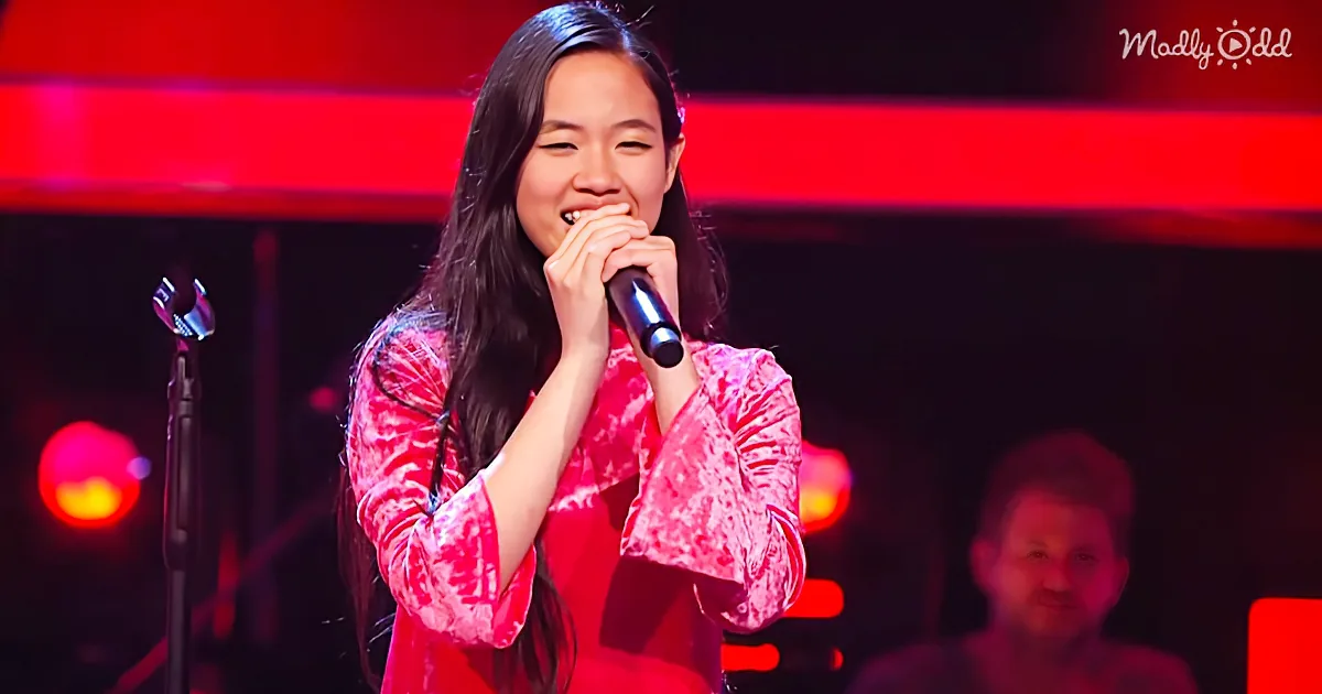 A woman in a red patterned shirt sings into a microphone on stage with red lighting.