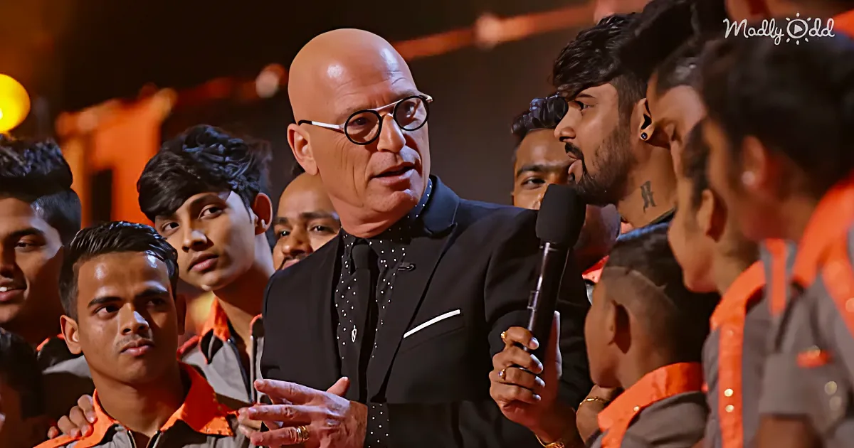 A man in a black suit and glasses speaks into a microphone surrounded by a group of people in orange uniforms.