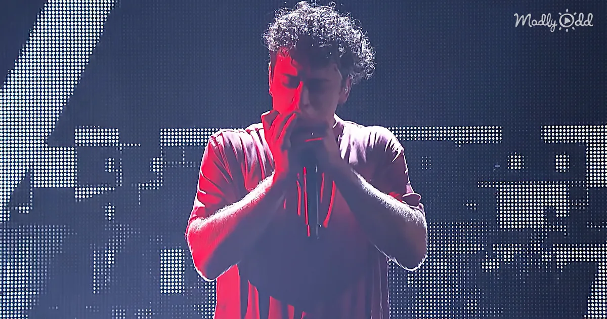 A person in a red shirt holds a microphone, illuminated by red light, against a digital background.