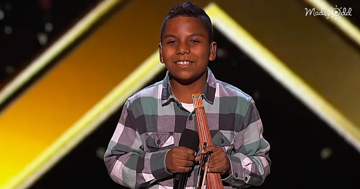A young boy in a plaid shirt holds a microphone on stage, smiling.