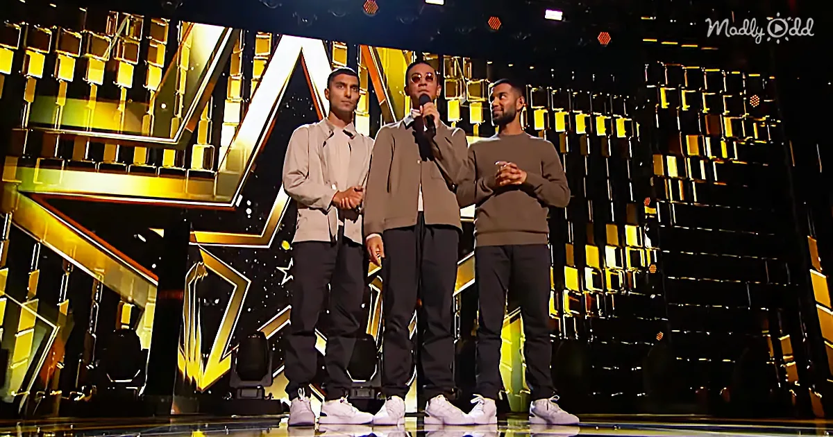 Three men stand on a stage with a microphone, wearing casual attire, against a vibrant, geometric backdrop.