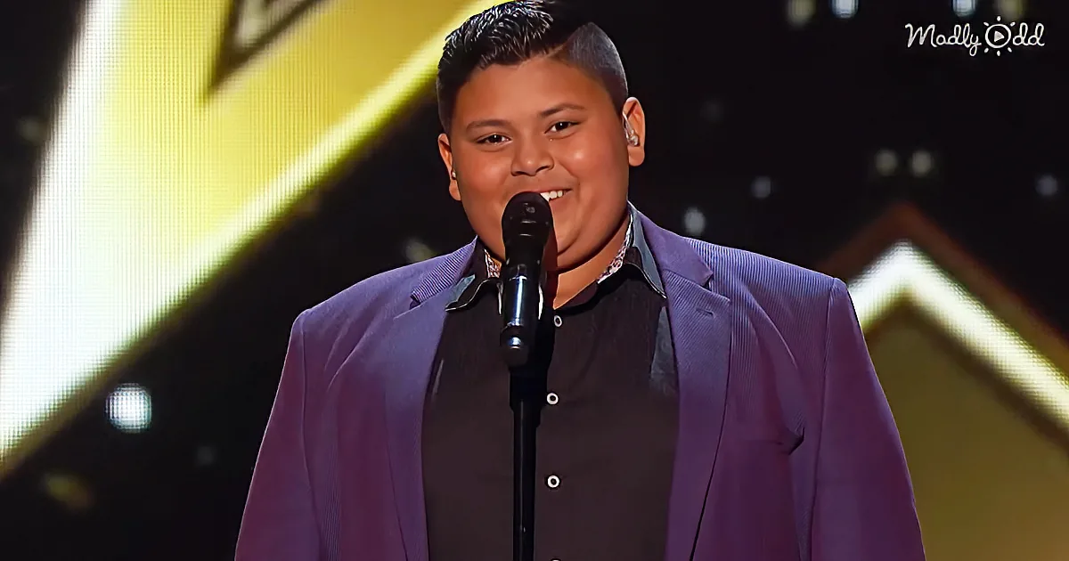 A young man in a purple suit stands on stage, smiling, with a microphone in front of him.