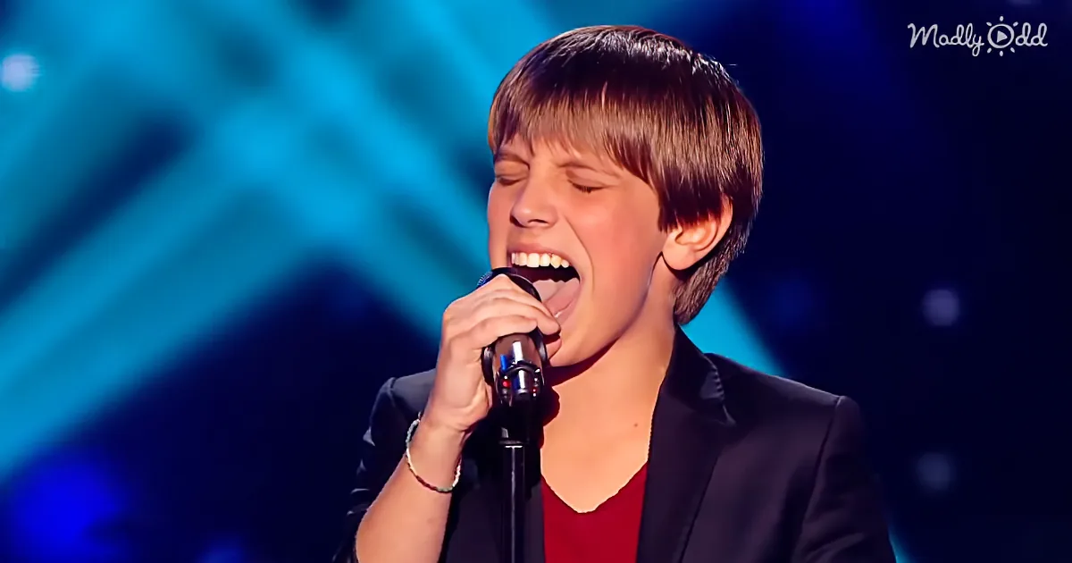 A young person in a black blazer sings passionately into a microphone on stage with blue lighting.