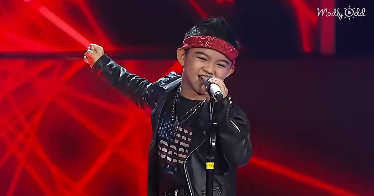 A young boy in a black leather jacket and red bandana sings into a microphone on stage with red lights.