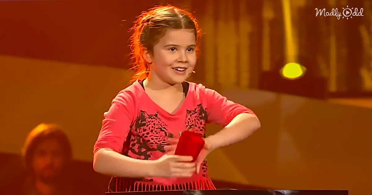 A young girl in a pink shirt smiles while holding a red object on stage.