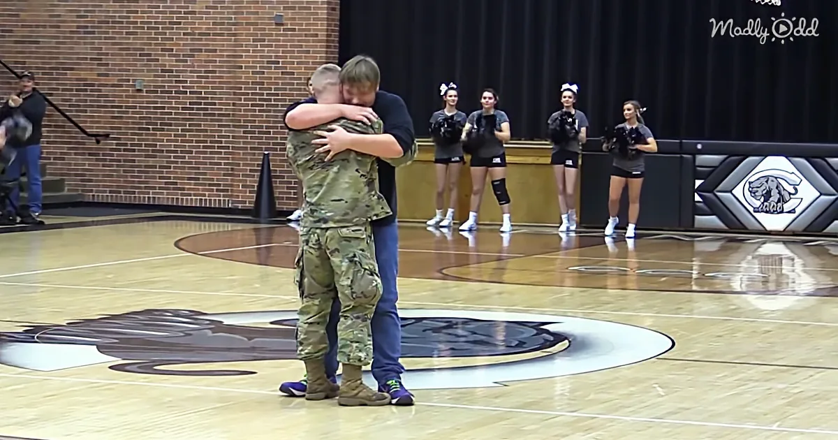 A soldier in camouflage hugs a person in a gymnasium, with cheerleaders and a photographer in the background.