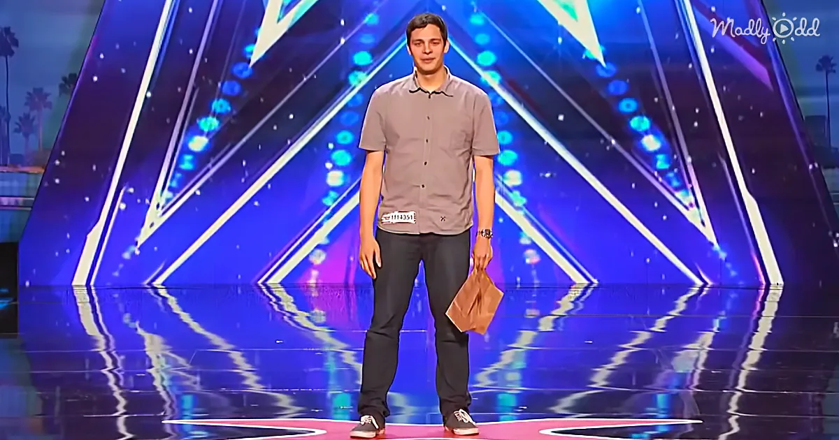 A man stands on a stage holding a brown paper bag, wearing a gray shirt and dark pants.