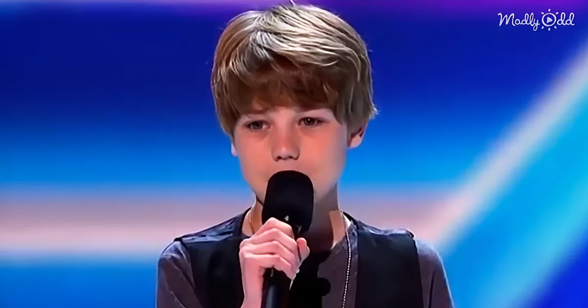 A young boy with short hair holds a microphone, standing against a blue background.