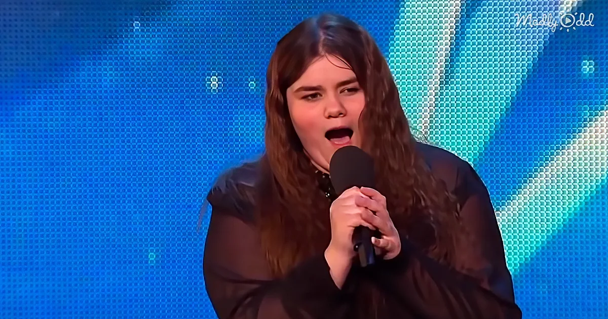 A person with long brown hair holds a microphone, singing on a stage with a blue background.