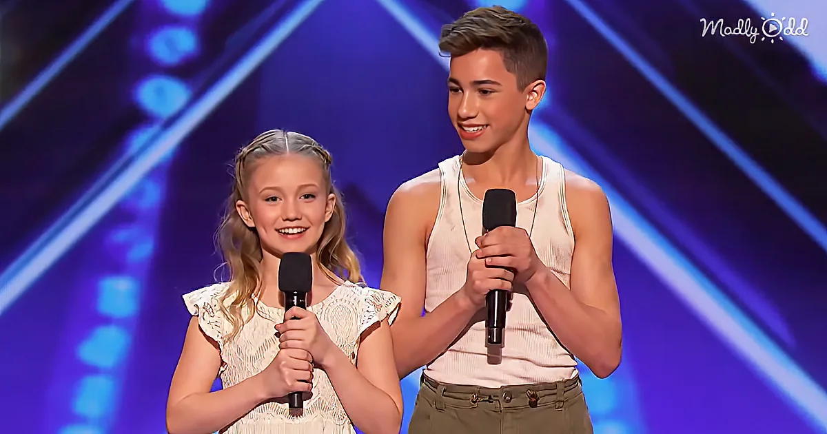 A young girl and boy hold microphones on stage, smiling, against a blue-lit backdrop.