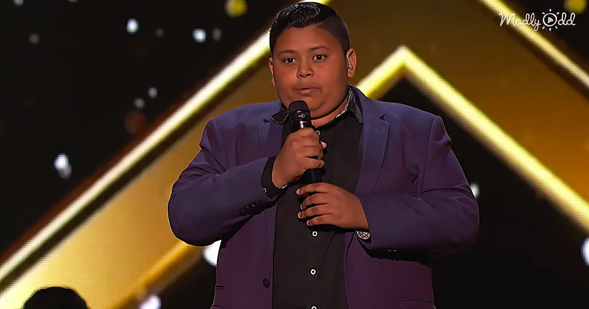A young man in a purple suit holds a microphone on stage, with a yellow and black background.