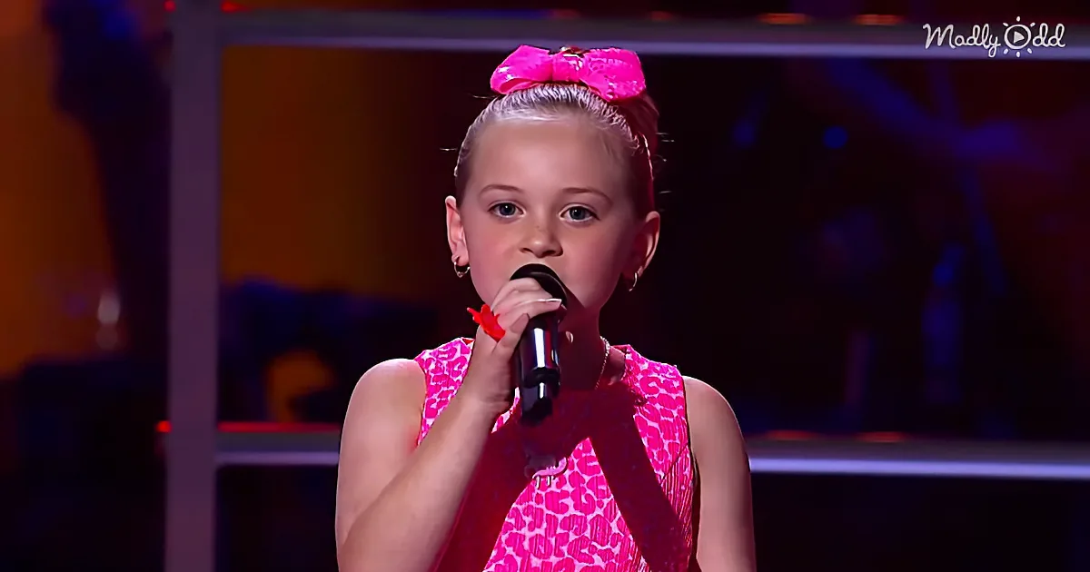 A young girl in a pink dress with a large pink bow sings into a microphone on stage.