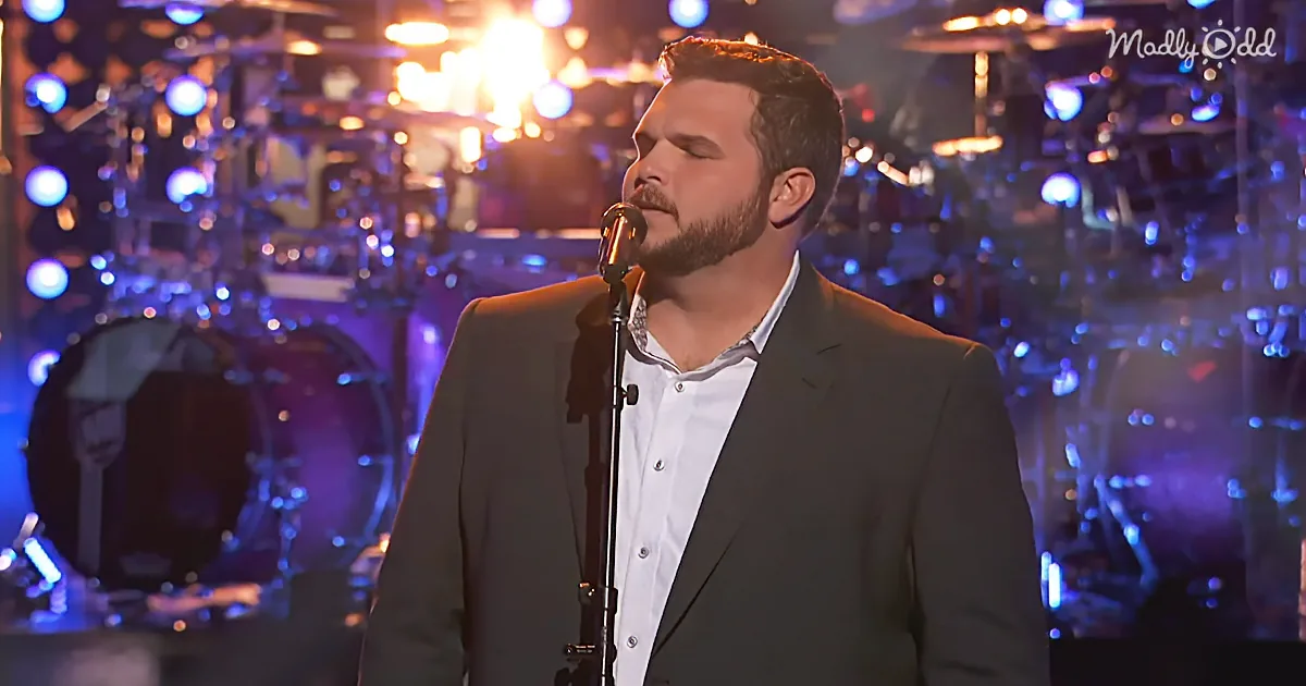 A man in a suit sings into a microphone on stage with a drum set and blue lights in the background.
