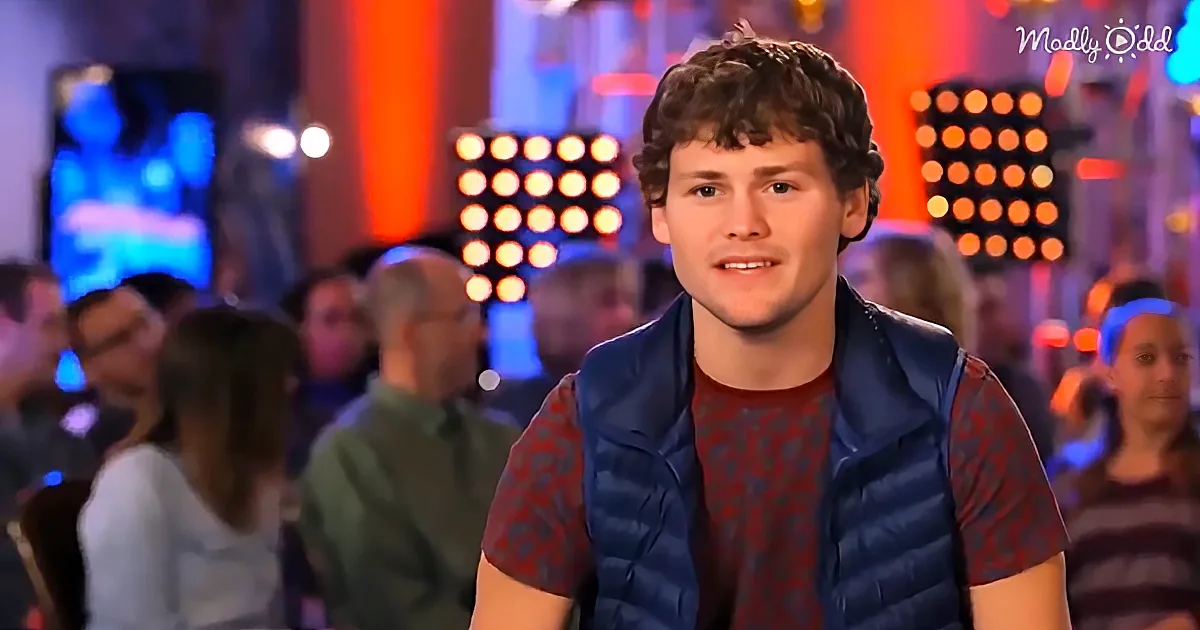 A man with curly hair, wearing a blue vest over a red patterned shirt, sits in a brightly lit room with a crowd and...