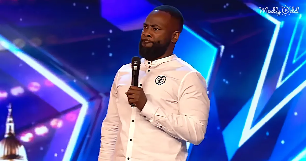 A man in a white shirt holds a microphone on stage with a blue geometric background.
