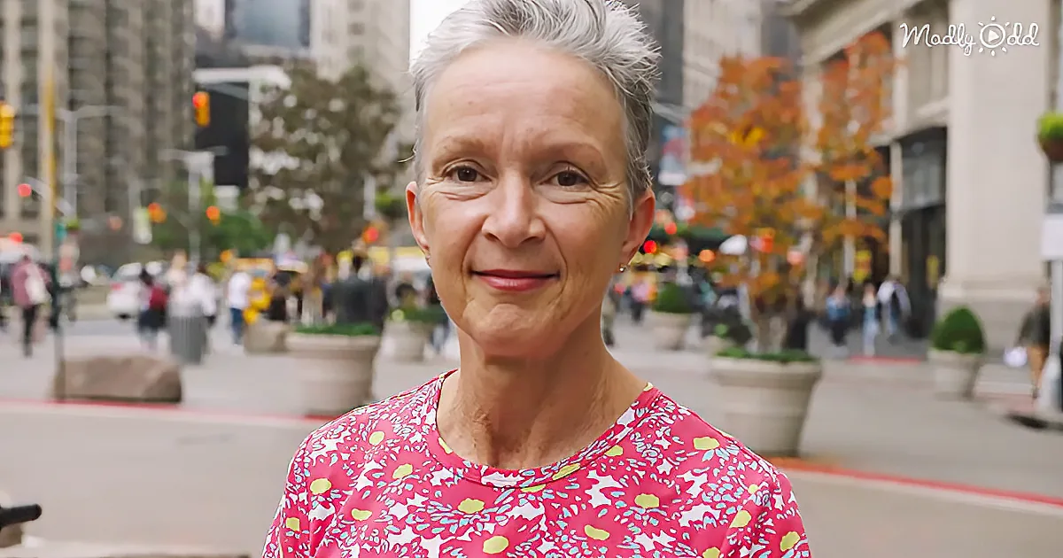 A woman with short gray hair smiles in a pink floral shirt on a busy city street with autumn trees and buildings in the...
