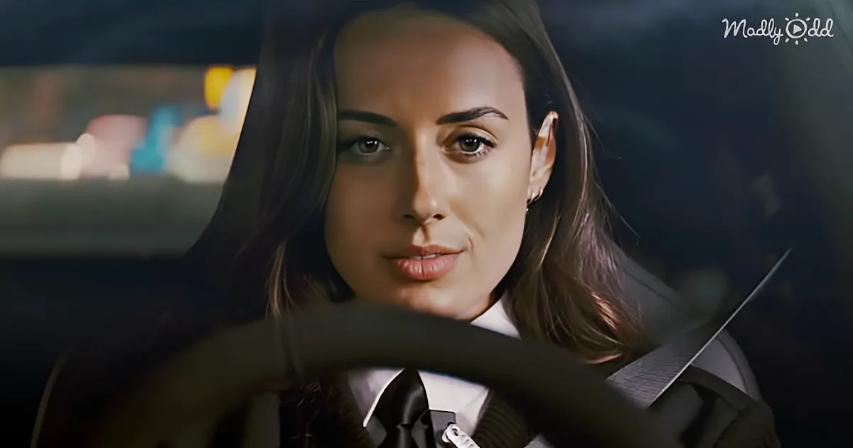 A woman with long brown hair, wearing a white shirt and tie, is driving a car at night.