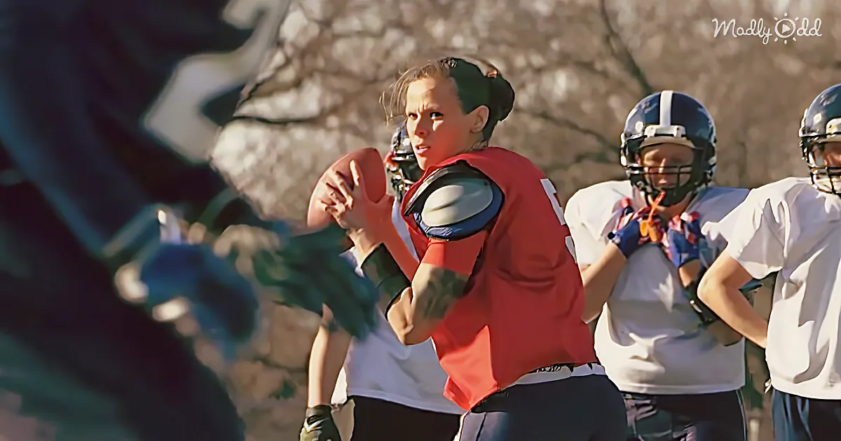 A young girl in a red jersey holds a football, preparing to throw, with teammates in white jerseys and helmets behind her.