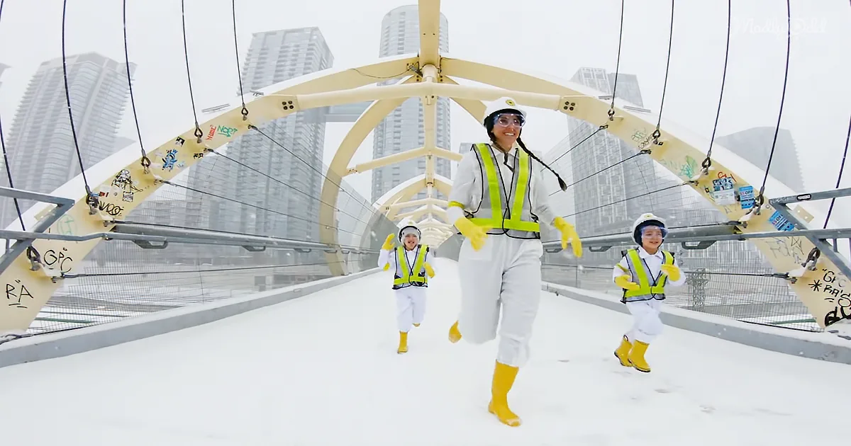 A woman and two children in safety gear run on a snowy bridge with tall buildings in the background.