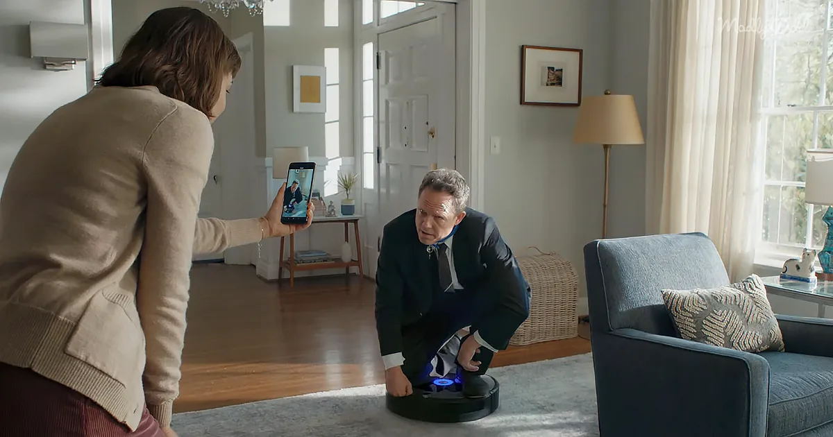A woman holds a phone showing a man, while another man kneels on a robotic vacuum cleaner in a cozy living room.