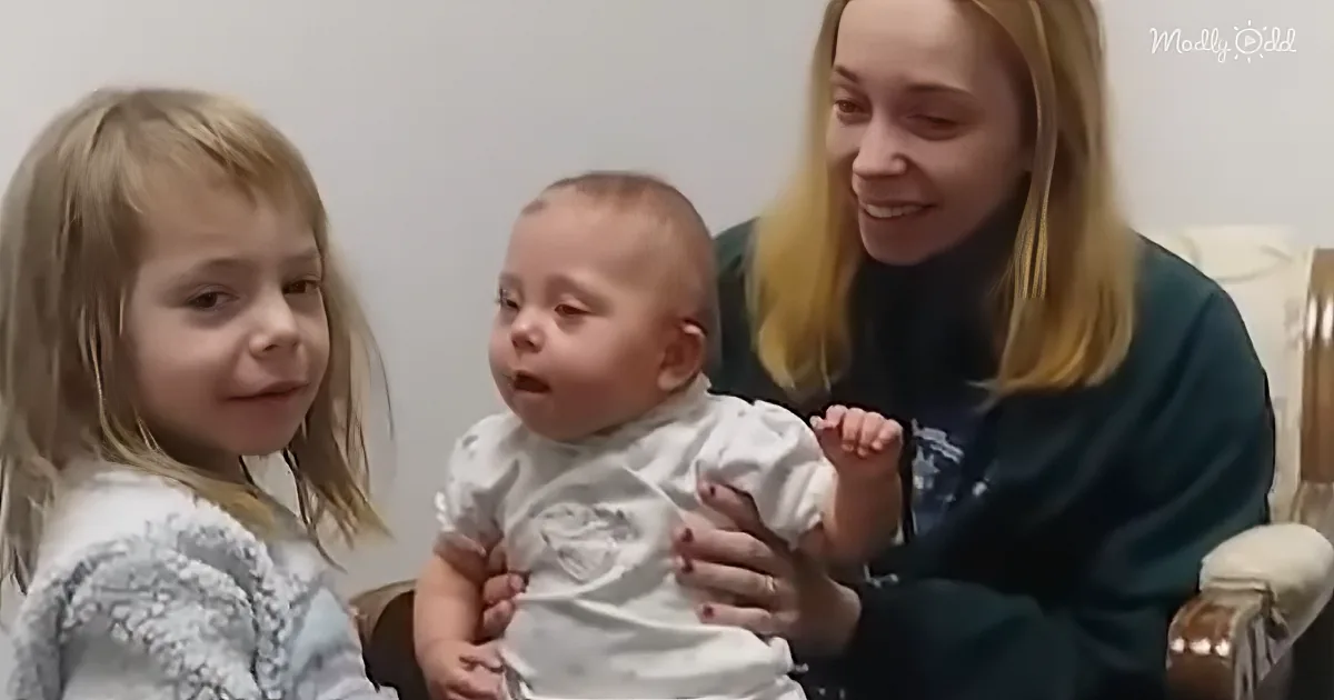 A woman holds a baby while a young girl looks on, both smiling.